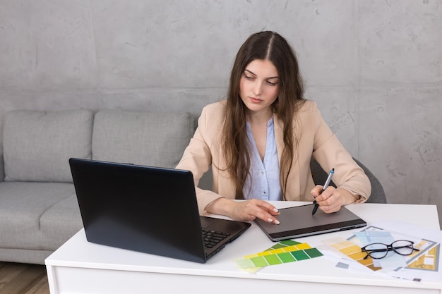 Photo a young designer girl sitting at a table draws on a tablet makes business calculations on the computer