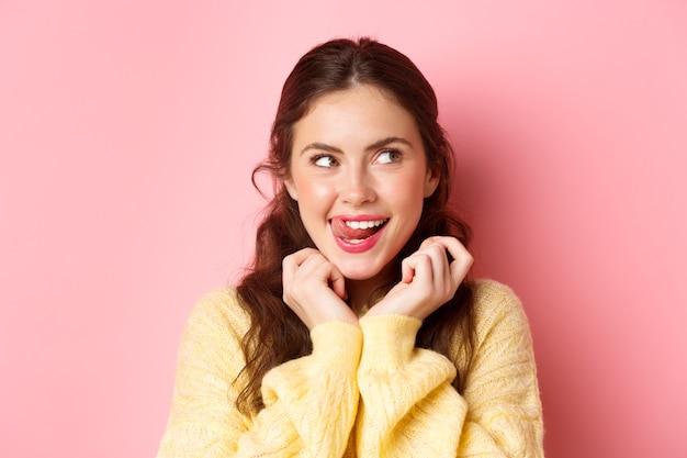 Young devious girl licking her lips and teeth, looking aside with thoughtful face, having interesting idea, thinking, standing against pink wall.