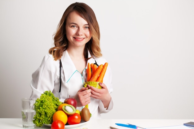 Young dietitian doctor at the consulting room at the table with fresh vegetables and fruits, working on a diet plan