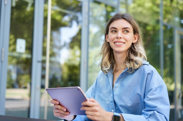 Young digital nomad woman with tablet working outdoors on fresh\
air smiling and looking away at pass