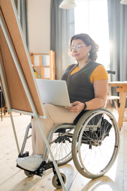 Young disable businesswoman looking at laptop display while preparing presentation for colleagues in front of whiteboard in modern office