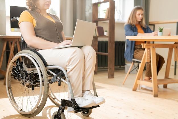 젊은 여성 사무원은 무릎을 꿇고 무릎을 꿇고 휠체어에 앉아 인터넷 서핑을 하는 동안 온라인 데이터를 살펴봅니다.
