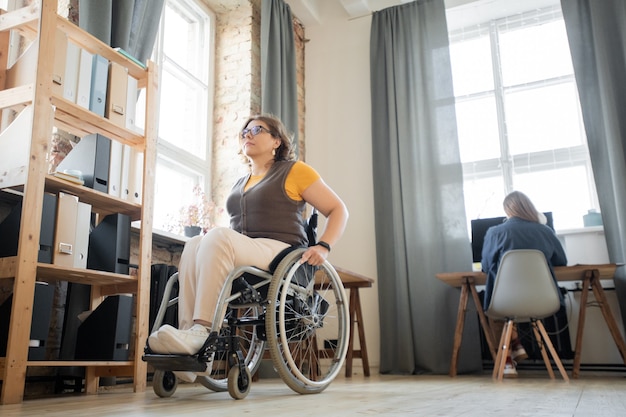 Молодая женщина-инвалид в инвалидном кресле движется к полке с бизнес-документами в современном офисе, где работает ее коллега