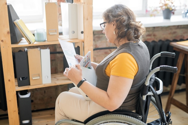 Young disable office secretary or accountant in wheelchair looking through financial papers while sitting by wooden shelves with group of folders