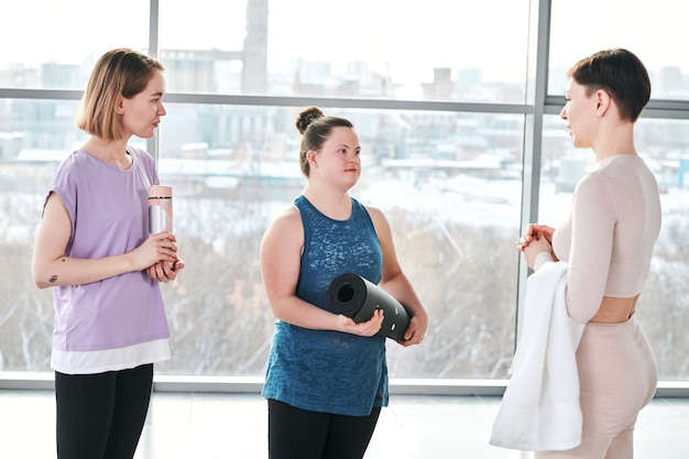 Молодая спортсменка-инвалид с черным свернутым ковриком консультируется с инструктором по фитнесу после тренировки со своей подругой, стоящей рядом