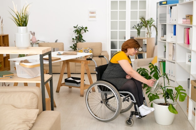 車椅子に座って、本棚のそばの床に立っている白い植木鉢で緑の国内植物に水をまくカジュアルウェアの若い障害のある女性