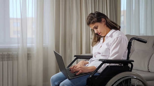 La giovane donna disabile dai capelli lunghi con paralisi cerebrale lavora online da remoto tramite laptop seduto su una sedia a rotelle vicino alla finestra nel soggiorno di casa