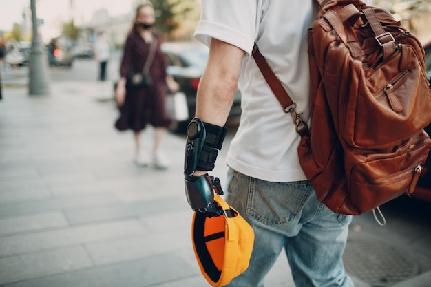 Молодой инвалид с искусственным протезом руки в повседневной одежде и рюкзаком гуляет по солнечной городской улице на открытом воздухе