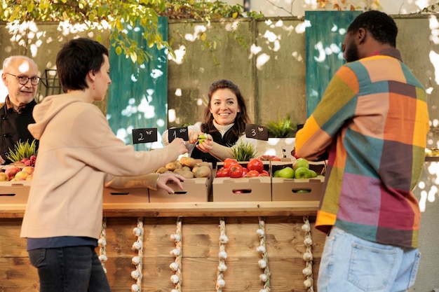 Young diverse family couple tasting different apple varieties while buying fresh organic fruits and vegetables at local farmers market. Friendly farm produce stand owner offering free samples