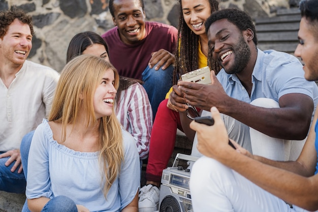 Young diverse people having fun listening music with boombox stereo outdoor in the city - Focus on african man holding cassette