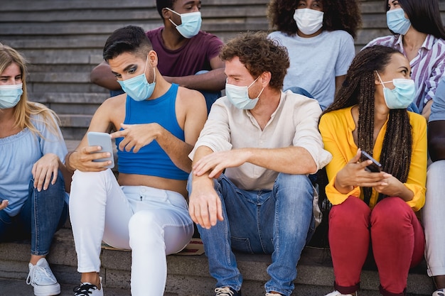 Young diverse people using mobile phones wearing safety mask outdoor in the city - Focus on gay man face