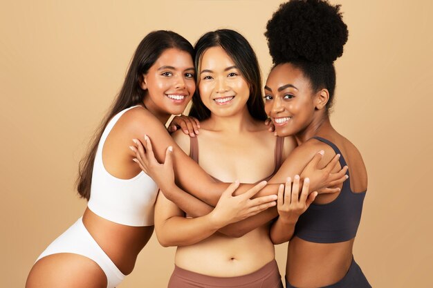 Young diverse women in underwear embracing on beige backdrop