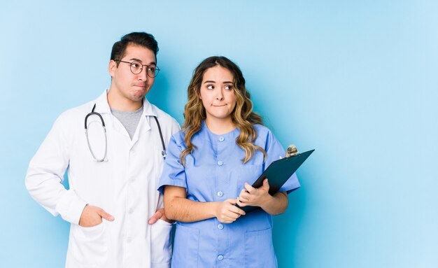 Young doctor couple posing in a blue wall isolated confused, feels doubtful and unsure.