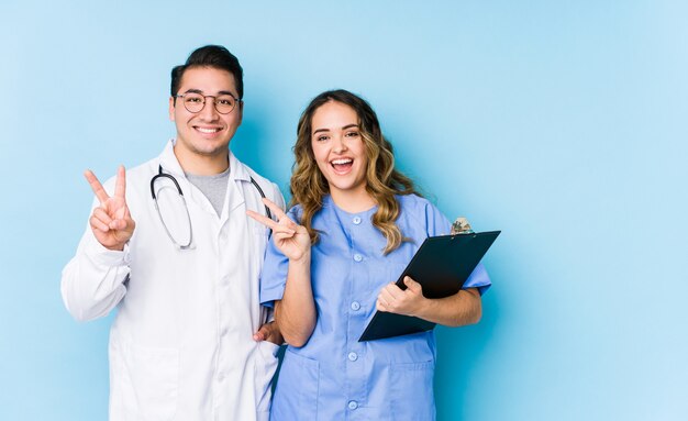 Young doctor couple posing in a blue wall isolated joyful and carefree showing a peace symbol with fingers