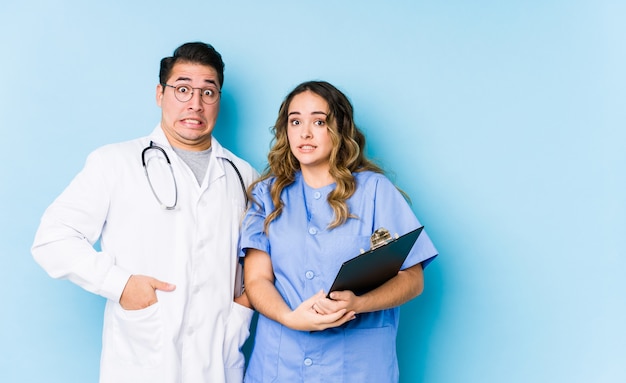 Young doctor couple posing in a blue wall isolated keeping eyes opened to find a success opportunity.