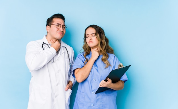 Young doctor couple posing in a blue wall isolated suffers pain in throat due a virus or infection.