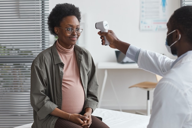 Young doctor examining the temperature of pregnant woman during her visit at hospital