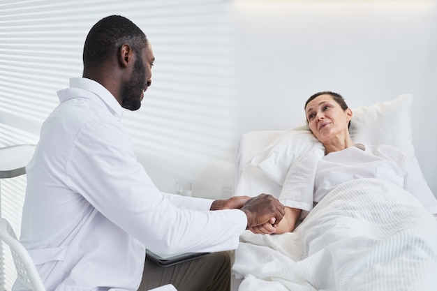 Young doctor holding hand of his patient and supporting her during illness