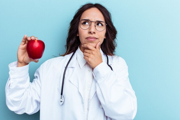 Young doctor latin woman holding an apple isolated on blue background