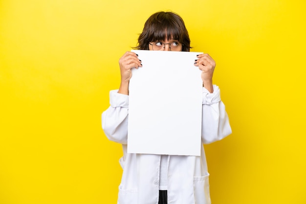 Young doctor latin woman isolated on yellow background holding an empty placard and hiding behind it