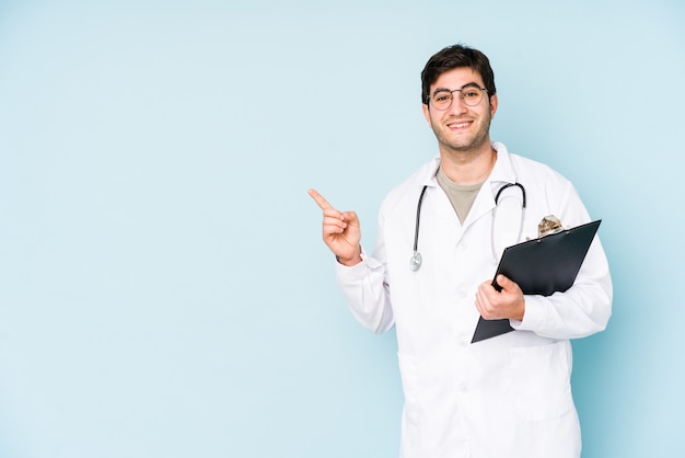 Young doctor man isolated on blue wall smiling and pointing aside, showing something at blank space.