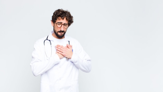 Young doctor man looking sad, hurt and heartbroken, holding both hands close to heart, crying and feeling depressed against copy space wall