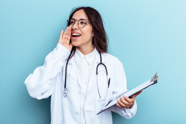 Young doctor mexican woman isolated on blue background shouting and holding palm near opened mouth.