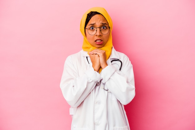 ピンクの背景に分離された若い医者のイスラム教徒の女性は怖くて恐れています。