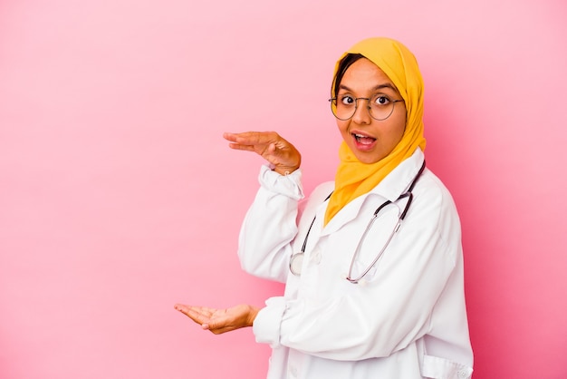 Young doctor muslim woman isolated on pink wall shocked and amazed holding a copy space between hands.