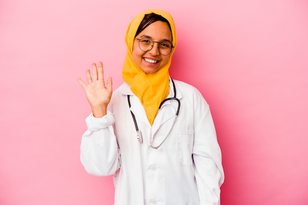 ピンクの壁に隔離された若い医者のイスラム教徒の女性は、指で5番を示す陽気な笑顔