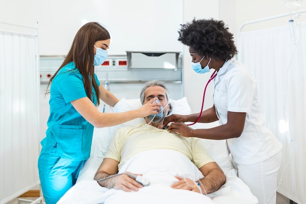 Young doctor and nurse wearing a surgical mask checking on a senior male patient wearing a positive pressure oxygen mask to aid breathing in a hospital bed