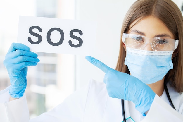 Young doctor in protective mask and gloves holding sos sign.