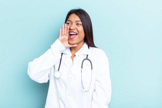 Young doctor Venezuelan woman isolated on blue background shouting and holding palm near opened mouth.