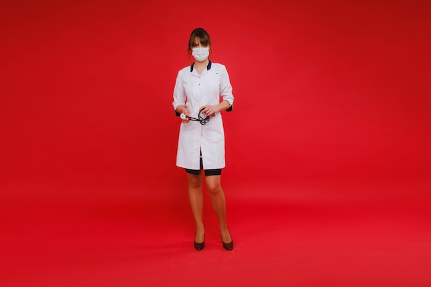 A young doctor in a white coat and medical mask stands on a red background