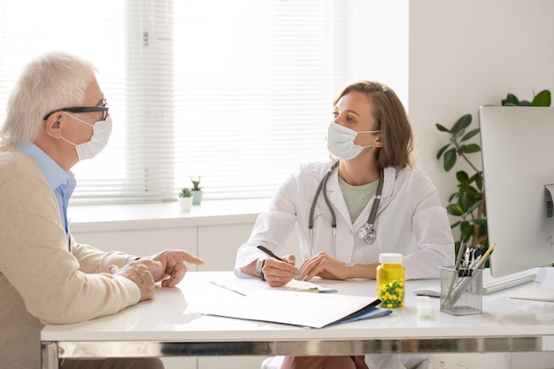 Young doctor in whitecoat and protective mask looking at senior man sitting in front of her, listening to him and making prescriptions