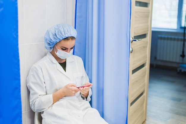 Young doctor with protective clothes sitting with her mobile phone during the break