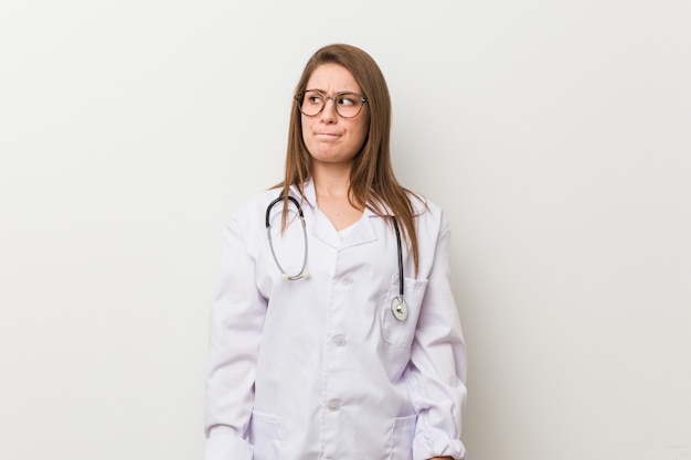 Young doctor woman against a white wall confused, feels doubtful and unsure.