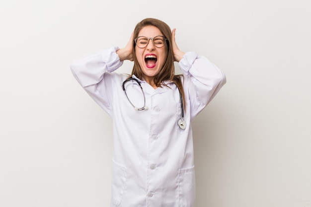 Young doctor woman against a white wall covering ears with hands trying not hearo loud sound.