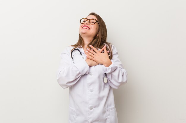 Young doctor woman against a white wall laughing keeping hands on heart