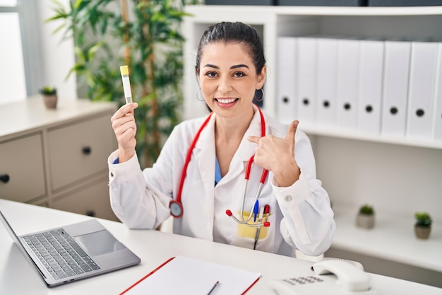 Young doctor woman holding blood sample pointing finger to one self smiling happy and proud