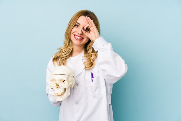 Young doctor woman holding a skull excited keeping ok gesture on eye.