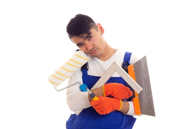 Young dolorous man with dark hair in white shirt and blue overall with orange gloves holding spatula, roll, ruler and screwdriver on white background in studio