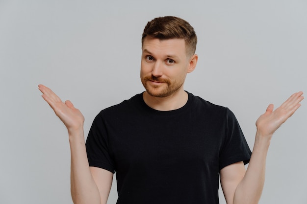 Young doubtful man in casual black t shirt cannot make decision and shrugging shoulders in uncertainty, looking at camera with facial expression of doubt and hesitation. Body language concept