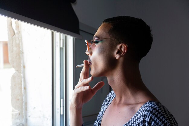 Photo young drag queen smoking in front of a window with make-up and dress