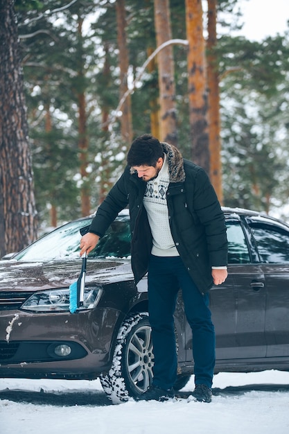 Молодой водитель мужчина зимой чистит фары автомобиля от снега