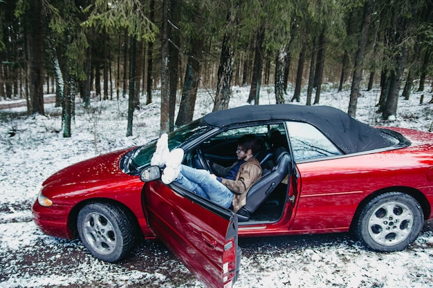 Giovane conducente in un'auto decappottabile rossa nella foresta invernale concetto di auto costosa