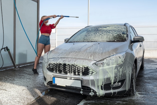 高圧ホースの若いドライバーの女性は、手動洗車で汚れをきれいにするために車に石鹸の泡を適用します