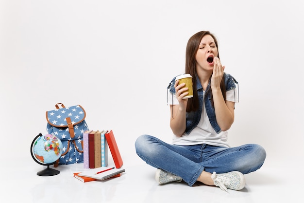 Young drowsy woman student holding paper cup with coffee or tea yawning want sleep sitting near globe, backpack, school books isolated