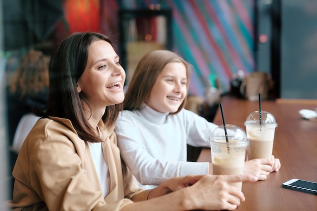 Young ecstatic female and her pretty daughter with milk cocktails laughing while looking through window in cafe