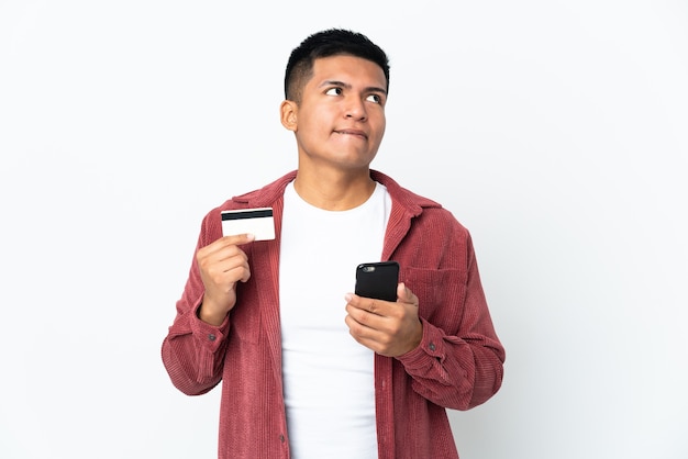 Young Ecuadorian man isolated on white wall buying with the mobile with a credit card while thinking